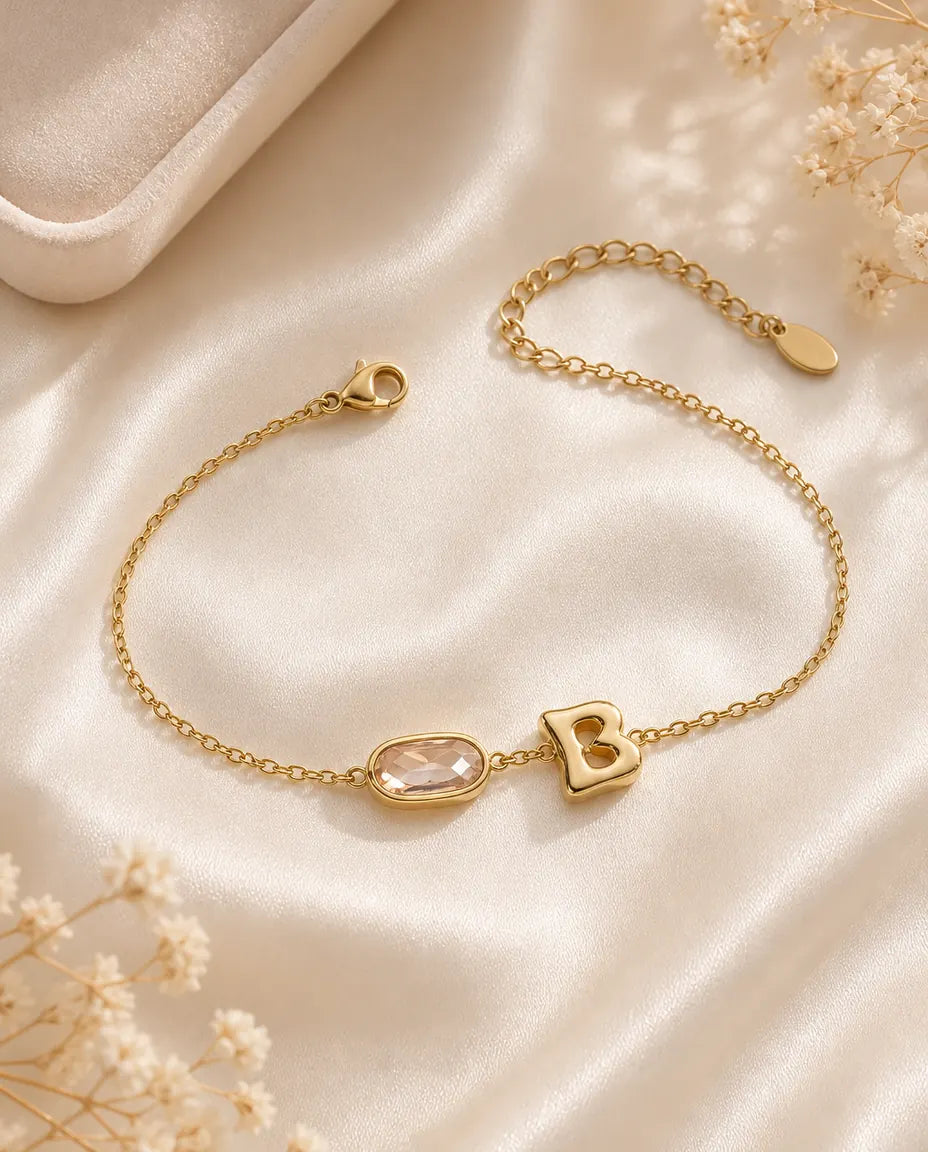 Personalised gold initial bracelet with an oval birthstone, flat lay on cream silk