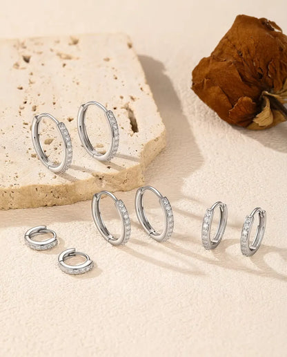 Moissanite Huggie Hoop Earrings in 925 Sterling Silver - all five sizes arranged on a cream surface by a travertine slab