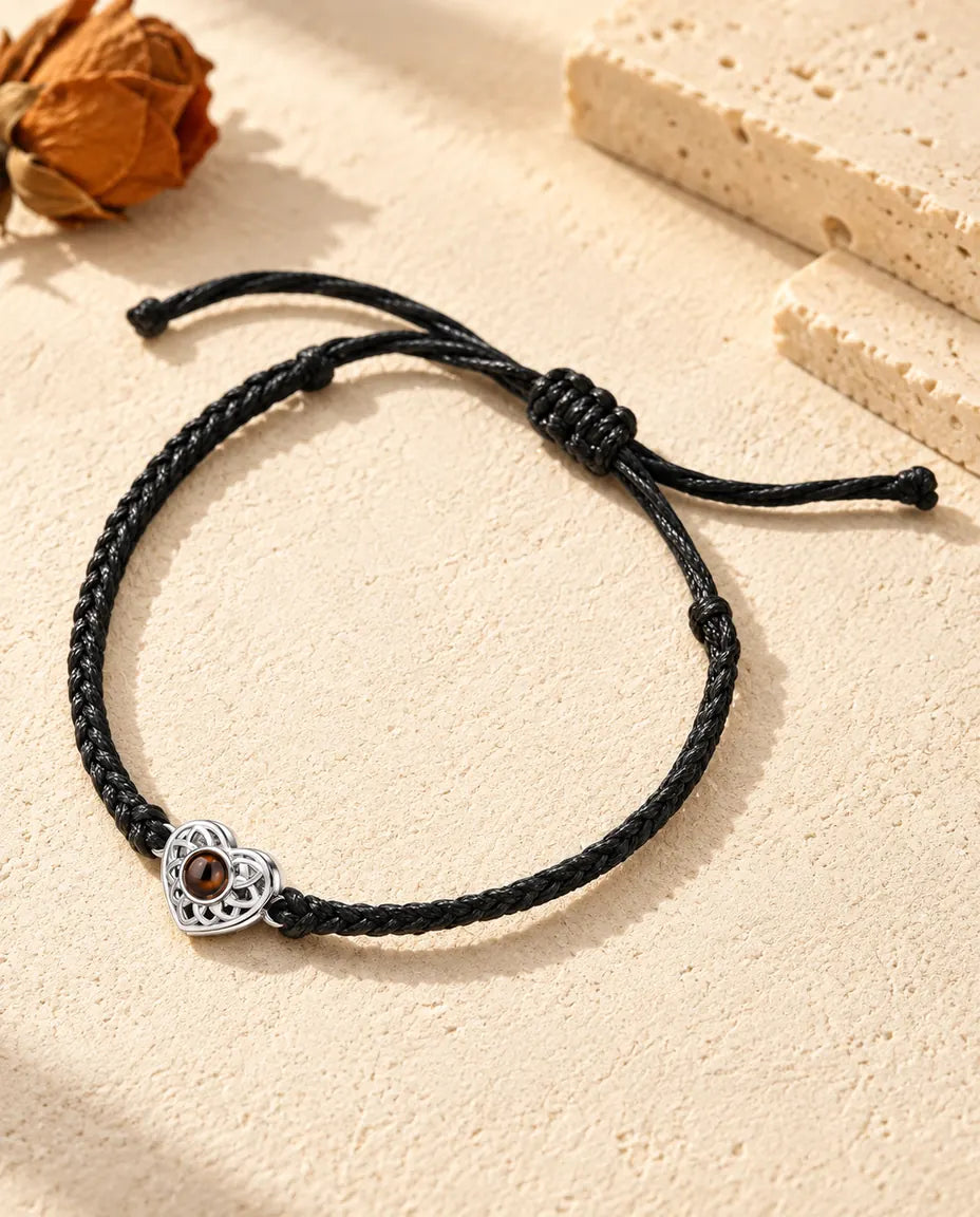 Personalised photo projection bracelet with a silver tone openwork heart charm on a black thin braided cord, lying on cream plaster