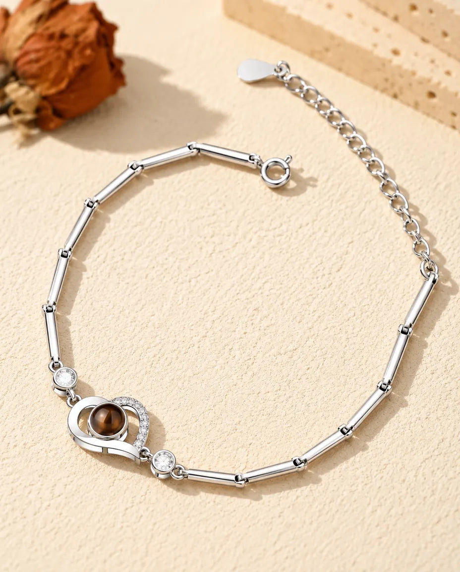 Personalised open heart photo projection bracelet in silver tone on a slim bar chain, lying on cream plaster