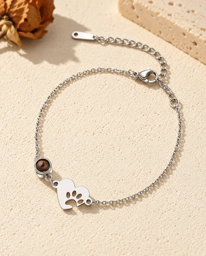 Personalised photo projection bracelet with a silver tone paw print heart tag and projection stone on a fine chain, lying on cream plaster