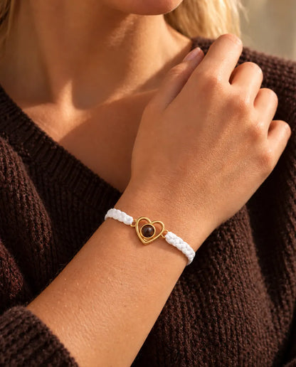 Close-up of the gold tone open heart charm on a white braided cord, fingers at the neckline