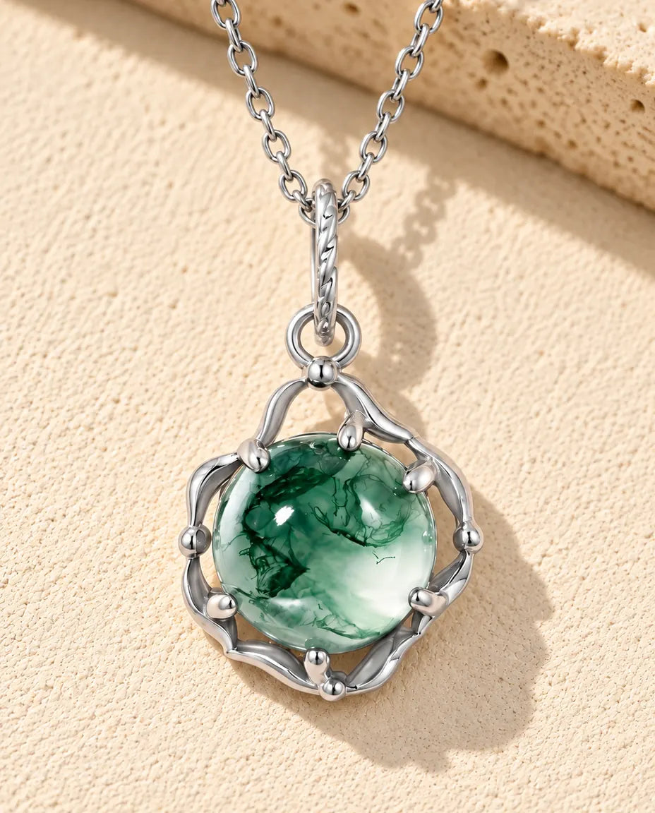 Round Moss Agate Cage Necklace in 925 Sterling Silver - lying on a cream textured surface beside a travertine block